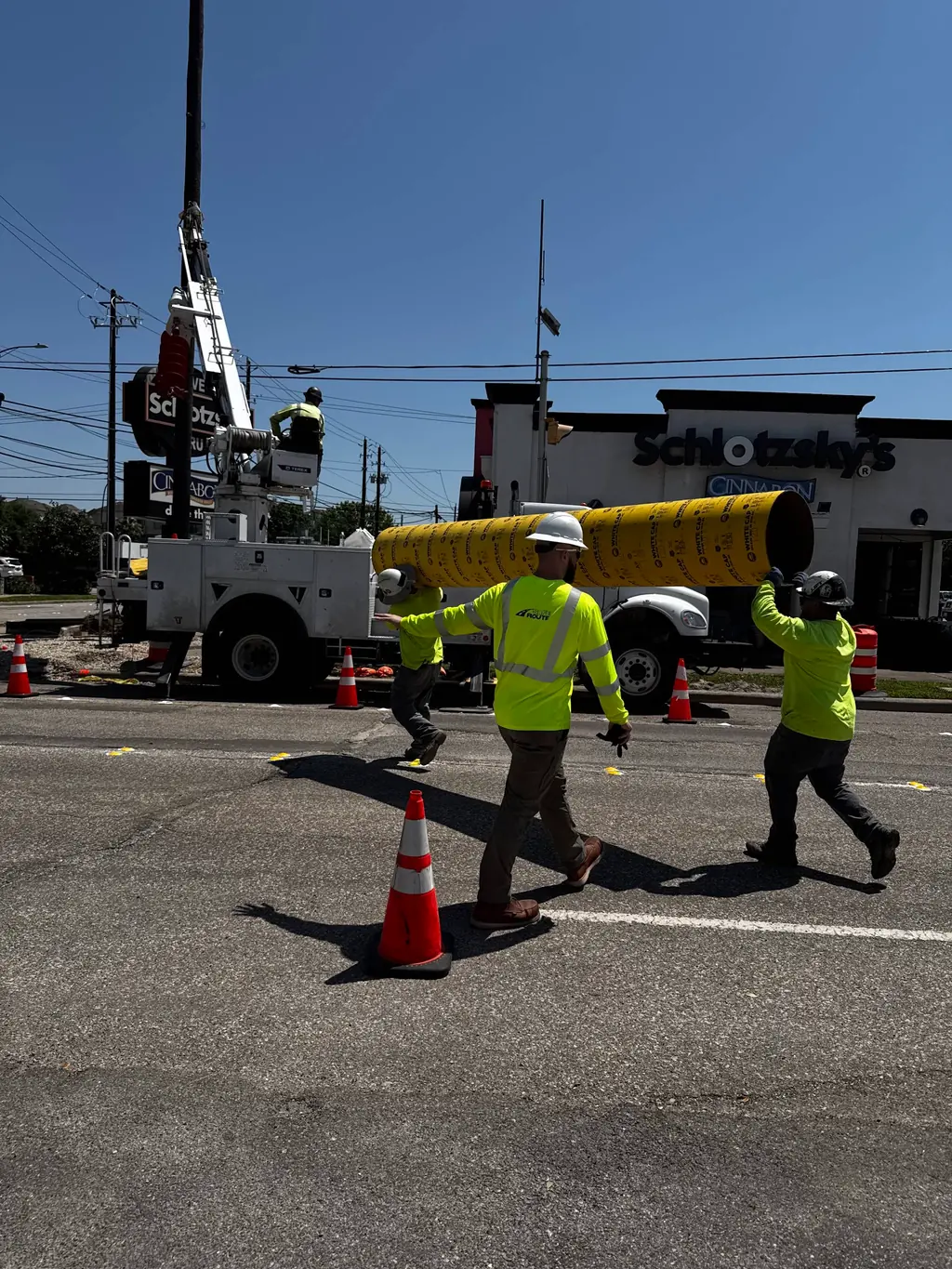 Workers carry a yellow pipe through a coned utility work area beside a bucket truck.
